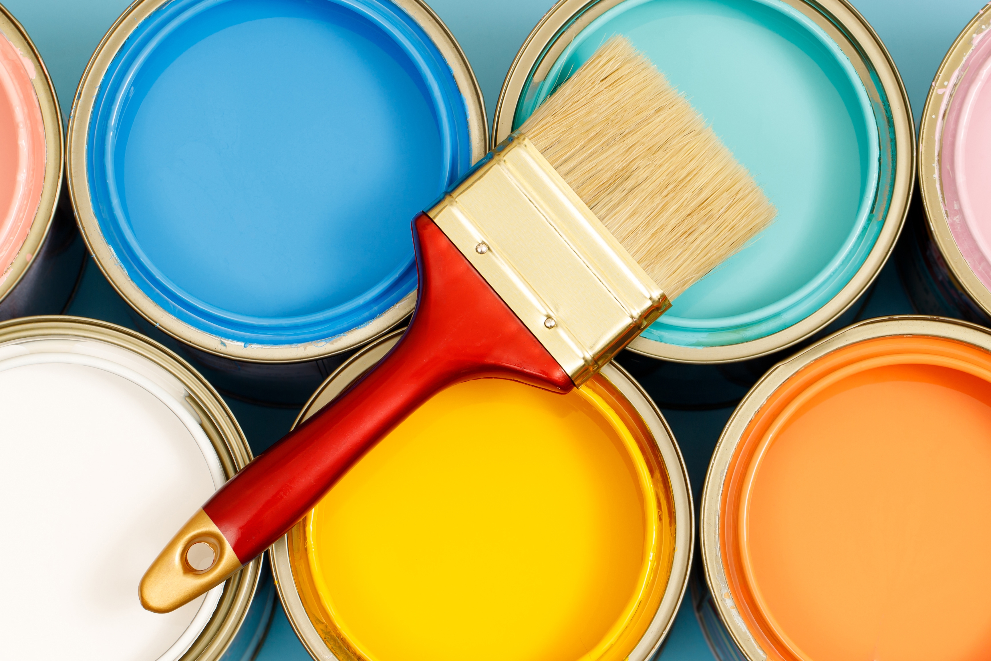 From above photographed paint cans in bright light blue, turquoise, white, yellow, and orange, as well as paint brushes.